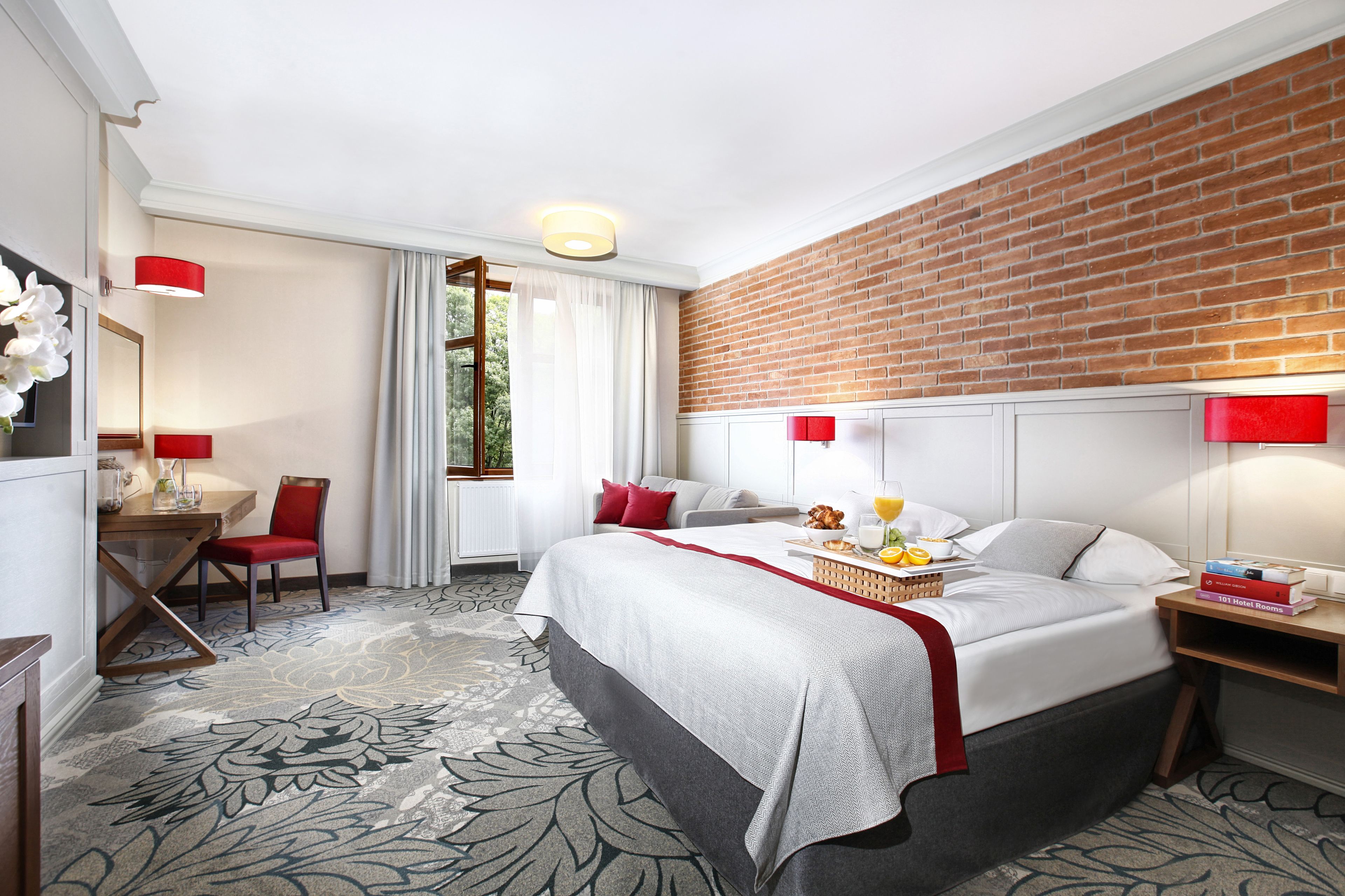 Une chambre d’hôtel avec un grand lit, un bureau avec une chaise, une petite table avec une lampe, un mur en briques et une fenêtre avec des rideaux.