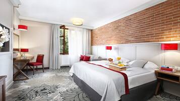 Une chambre d’hôtel avec un grand lit, un bureau avec une chaise, une petite table avec une lampe, un mur en briques et une fenêtre avec des rideaux.
