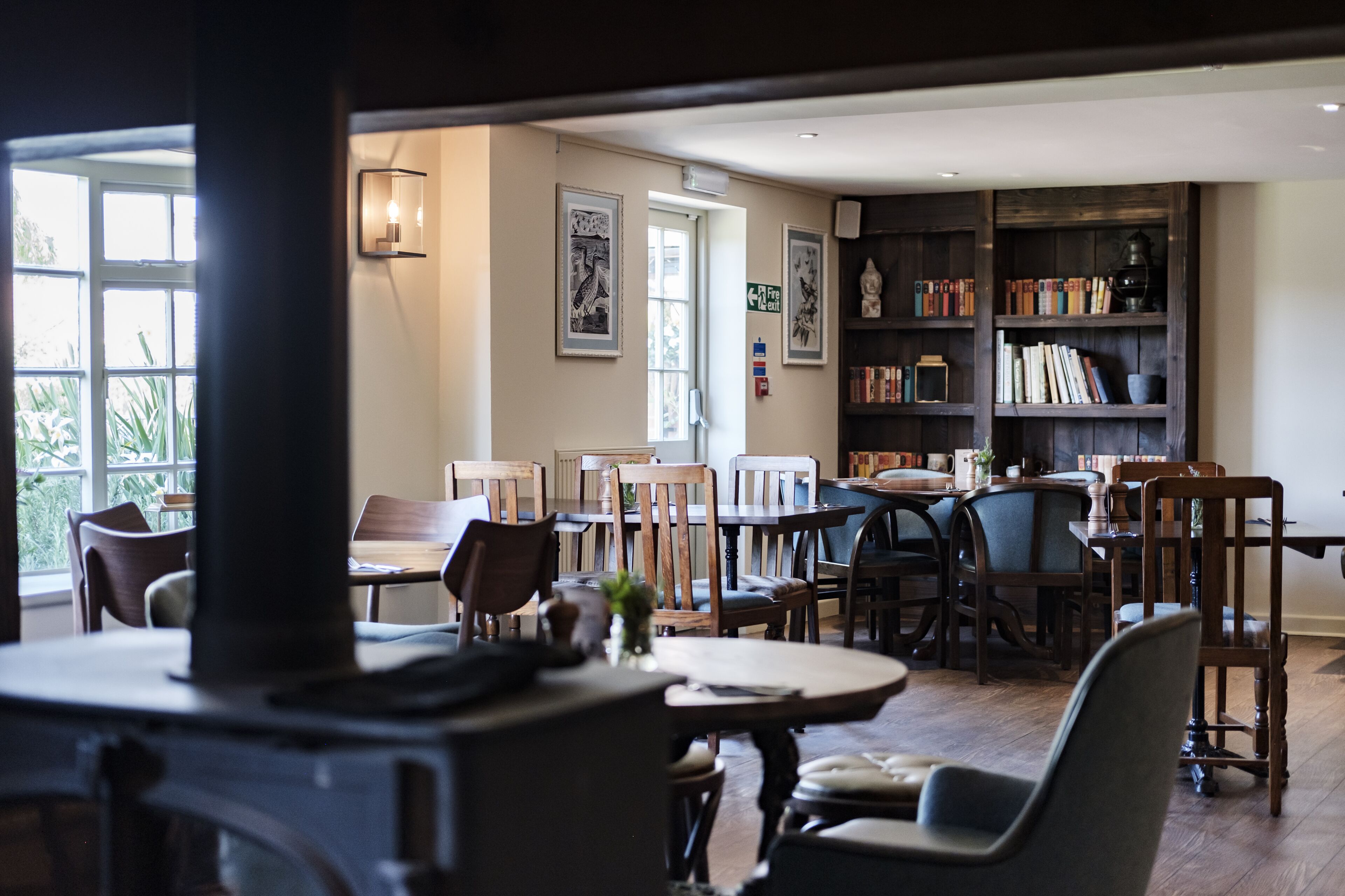 Een gezellig eetgedeelte met houten tafels en stoelen, een boekenkast en een groot raam met natuurlijk licht.