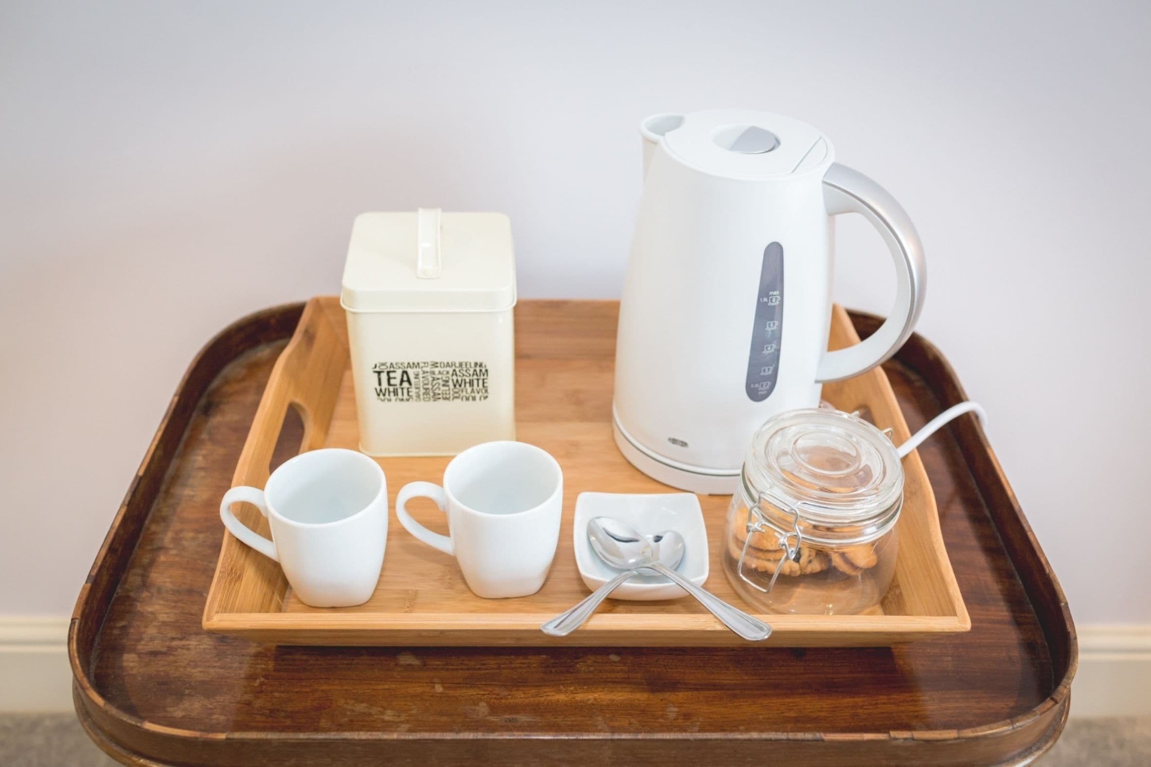 A tray with a white electric kettle, two white cups, a white box labeled "TEA", a small glass jar with cookies, and a spoon on a wooden tray.