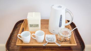A tray with a white electric kettle, two white cups, a white box labeled "TEA", a small glass jar with cookies, and a spoon on a wooden tray.