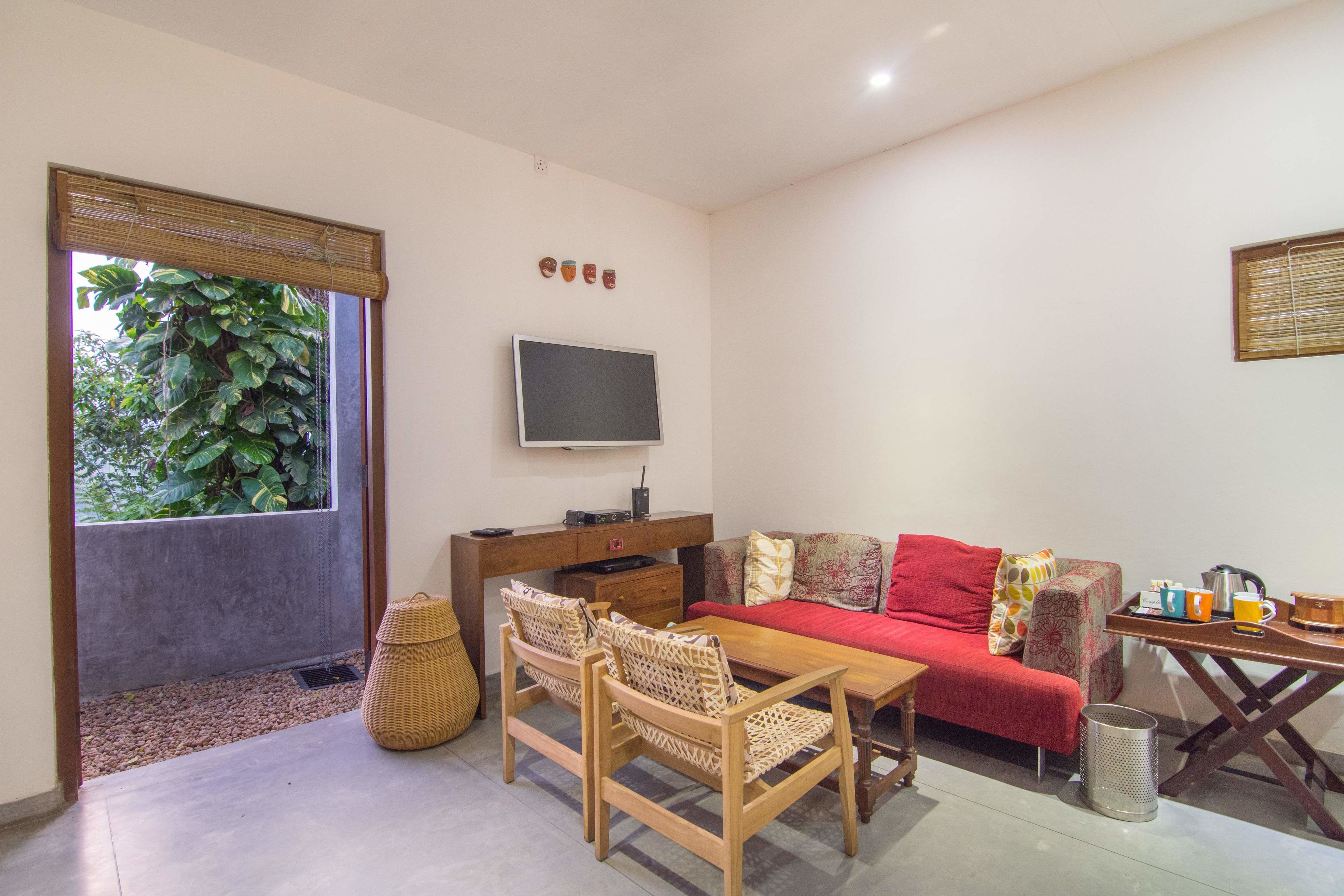 A room with a wooden table, two chairs, a sofa, a TV, and a window with a view of greenery.
