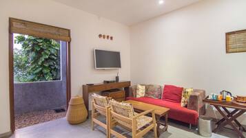 A room with a wooden table, two chairs, a sofa, a TV, and a window with a view of greenery.