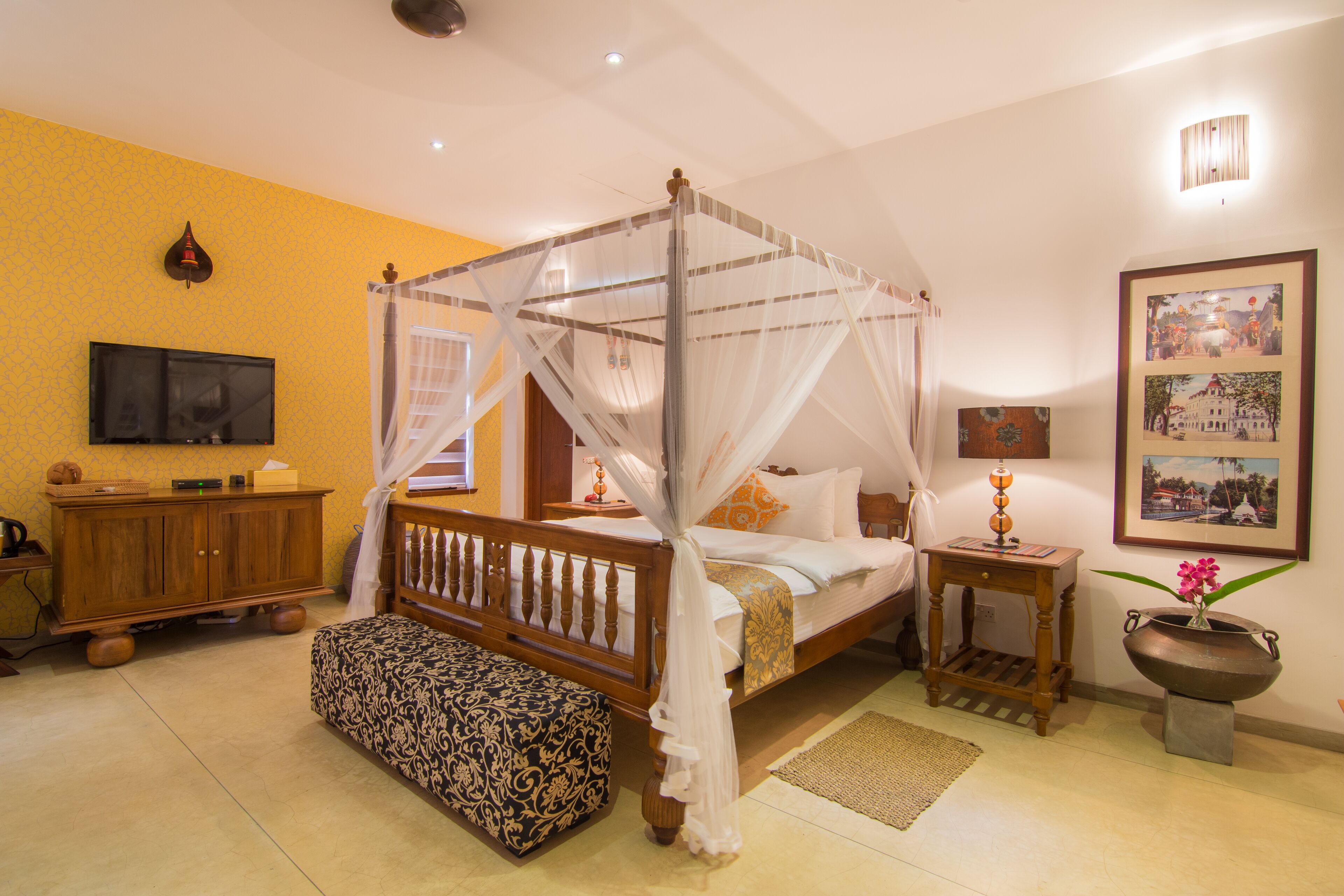 A four-poster bed with a canopy, a wooden bench, a bedside table with a lamp, and a wall-mounted TV.