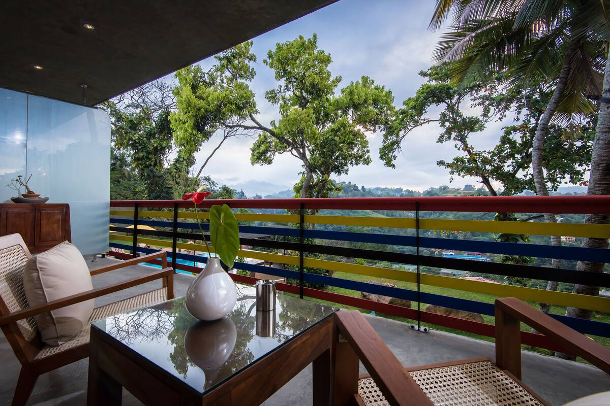 A balcony with a glass table, wooden chairs, and a view of trees and mountains.