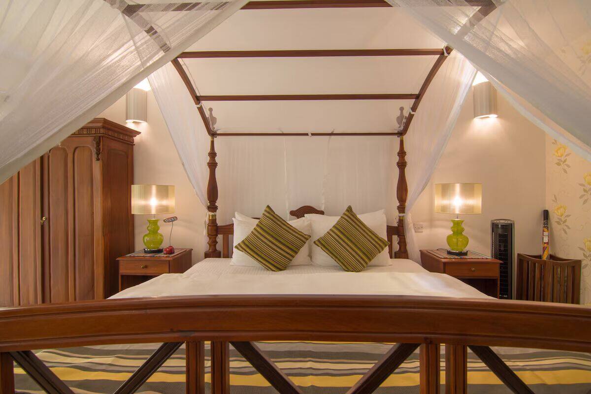 A four-poster canopy bed with white bedding and green striped pillows, wooden nightstands with lamps, and a wooden wardrobe.