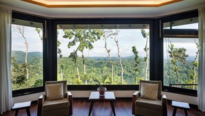 A room with large windows offering a view of a forested landscape. There are two chairs and a small table in the room. - The Elephant Stables (Kandy)