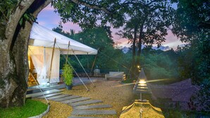 A tented accommodation with a stone pathway leading to a seating area, surrounded by trees and a view of a traditional building. - The Elephant Stables (Kandy)