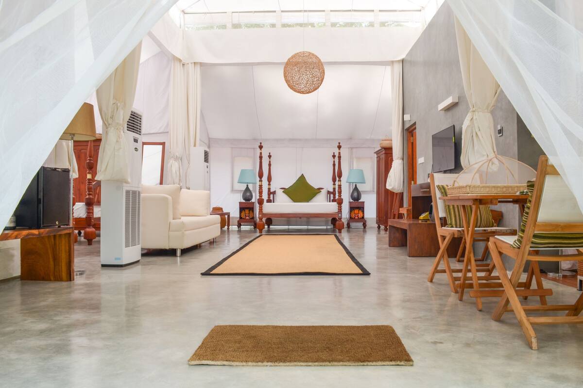 A spacious living room with a large sofa, a coffee table, and a TV. There is a large window with curtains, a ceiling fan, and a decorative hanging light.