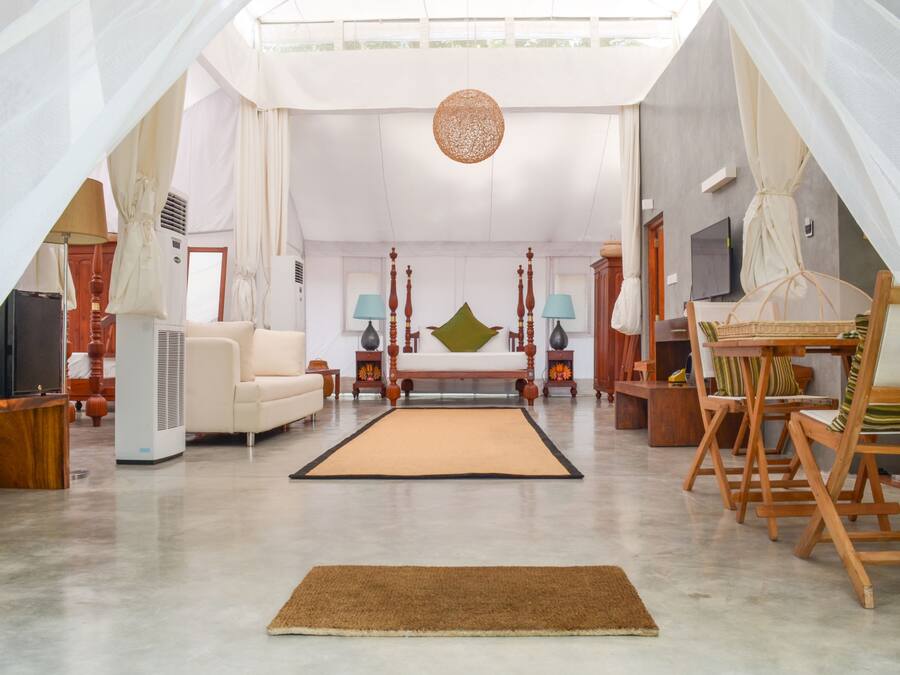 A spacious living room with a large sofa, a coffee table, and a TV. There is a large window with curtains, a ceiling fan, and a decorative hanging light.