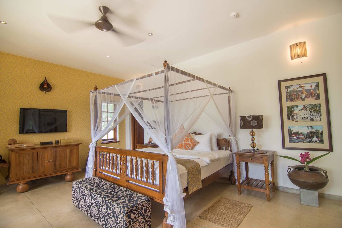 A bedroom with a four-poster bed, a wooden nightstand, a television, and a wall-mounted lamp.