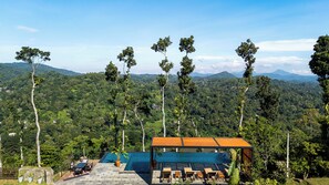 Un área de piscina con terraza de madera, sillas de descanso y vistas a un bosque exuberante y montañas al fondo.