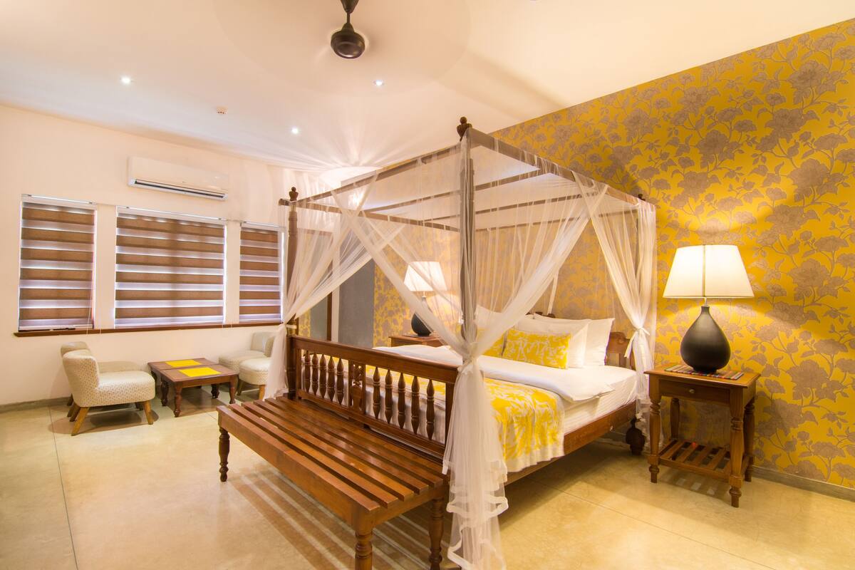 A four-poster canopy bed with a bedside table and lamp, a wooden bench, two armchairs, and a window with blinds.