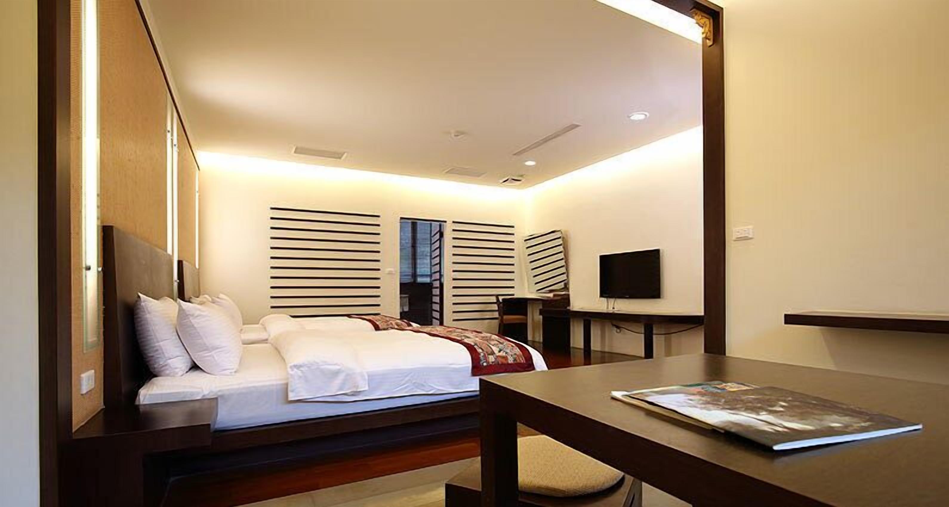 A modern hotel room with a bed, bedside table, TV, and a window with blinds.