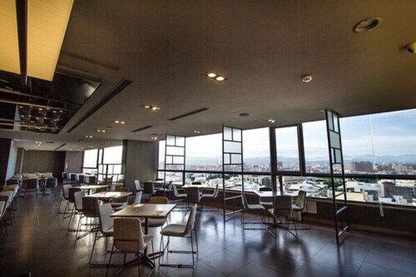 Un espacio interior amplio con mesas y sillas dispuestas para comer o reuniones, ventanas grandes que ofrecen vistas a la ciudad y un diseño de techo moderno.