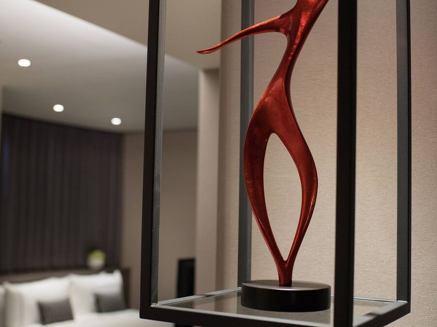 Quarto de hotel moderno com cama, mesa de cabeceira e uma escultura decorativa vermelha numa moldura preta.