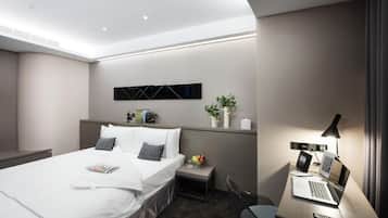 A modern hotel room with a neatly made bed, a desk with a laptop, a remote control, and a small plant.