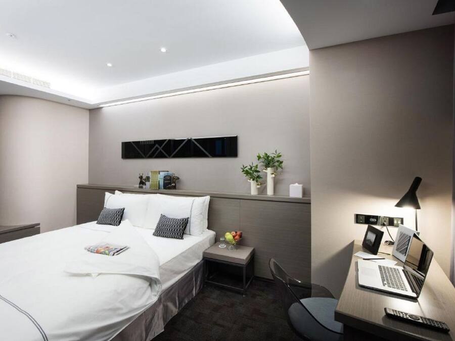 A modern hotel room with a neatly made bed, a desk with a laptop, a remote control, and a small plant.