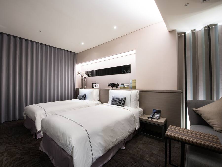 A hotel room with two beds, a bedside table with a phone, a small table with a vase, and a large window with curtains.