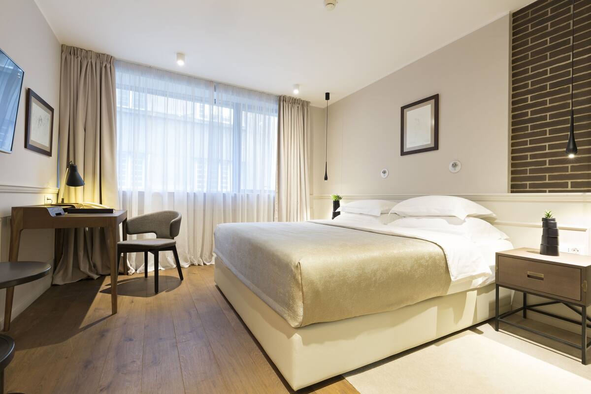A modern hotel room with a large bed, a desk with a chair, a bedside table with a lamp, and a view of the city through sheer curtains.