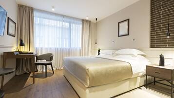 A modern hotel room with a large bed, a desk with a chair, a bedside table with a lamp, and a view of the city through sheer curtains.