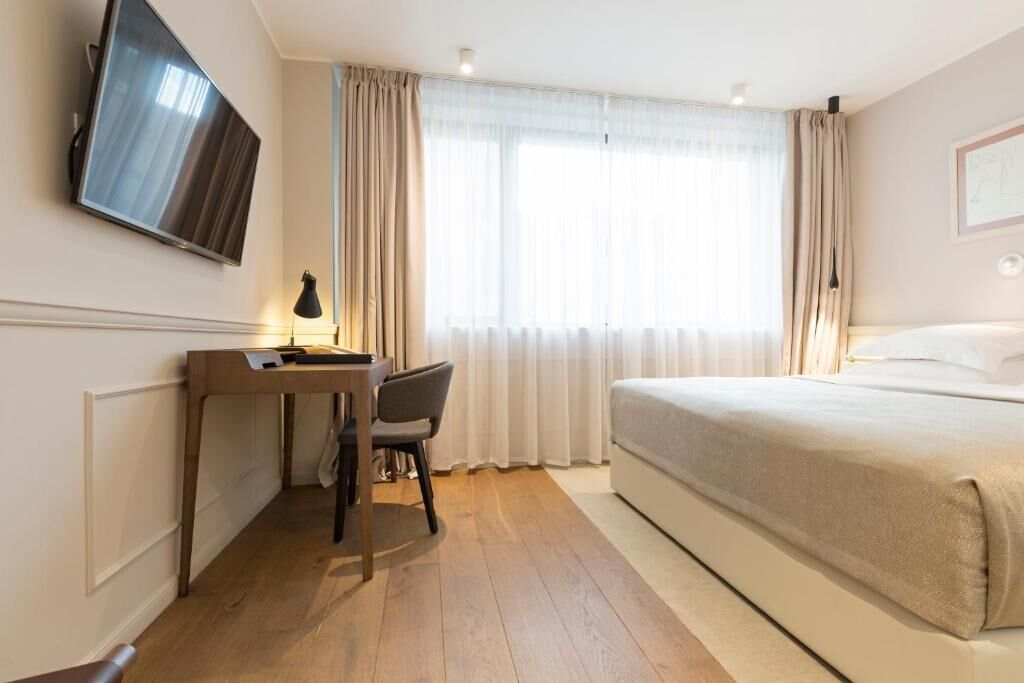 Habitación de hotel con cama, escritorio con silla, televisor montado en la pared y ventana con cortinas.