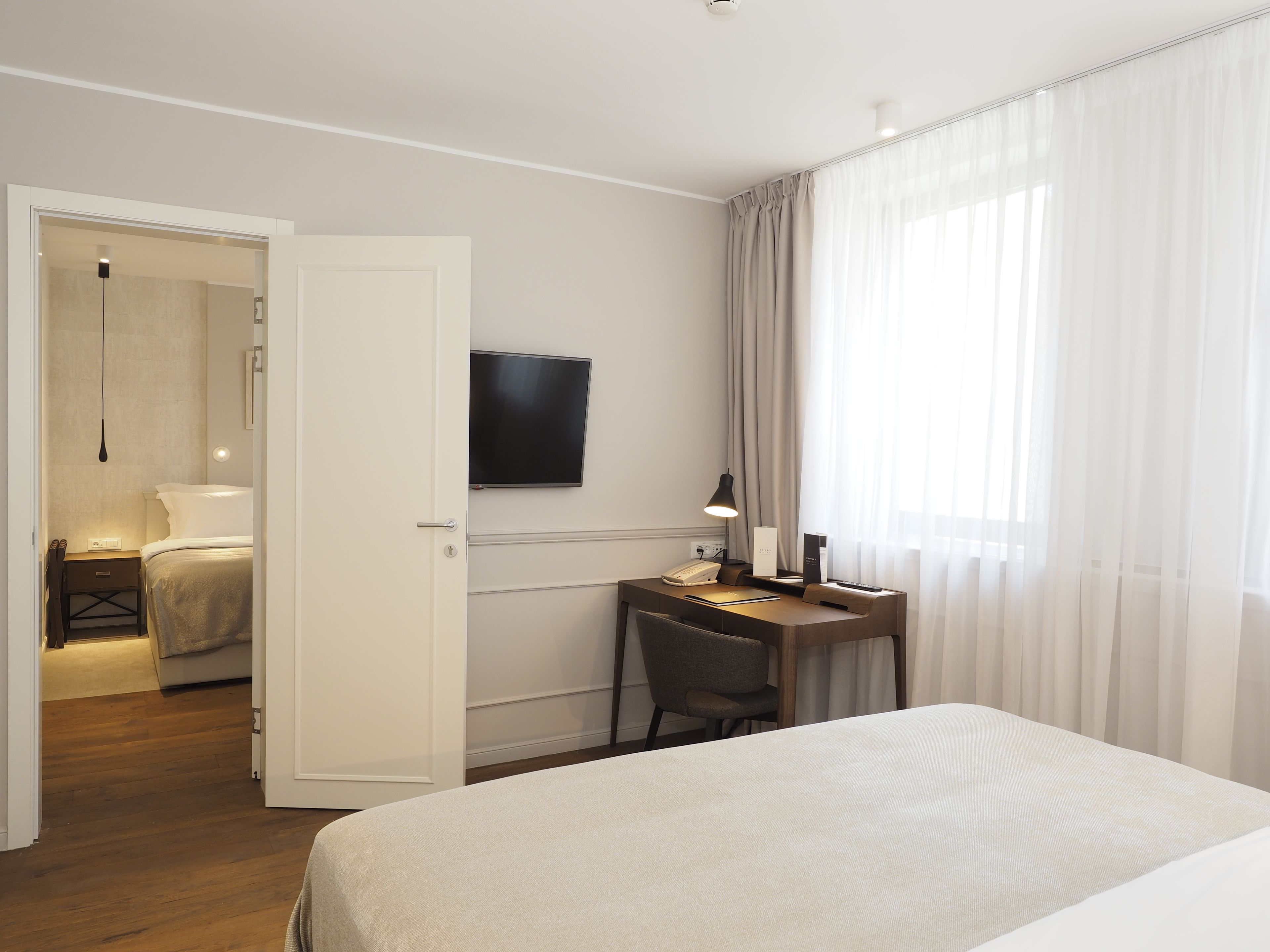 A hotel room with a bed, a desk with a chair, a TV mounted on the wall, and a window with curtains.