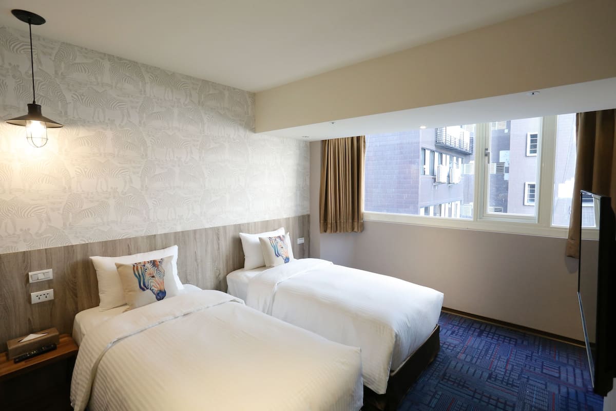 A hotel room with two beds, a patterned wall, a window with curtains, and a view of buildings outside.