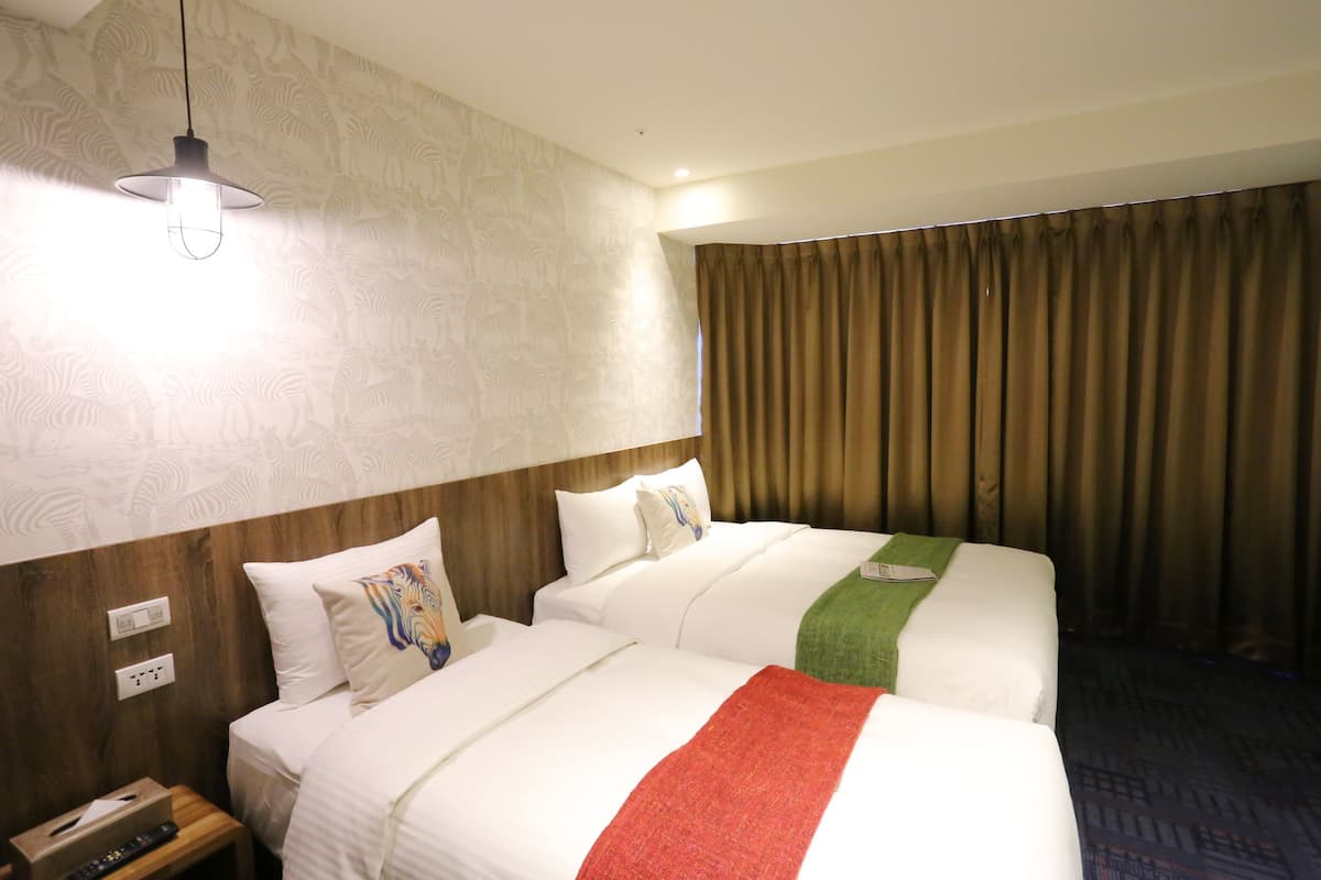 A hotel room with two beds, a wooden headboard, a nightstand with a remote control, and a window with curtains.