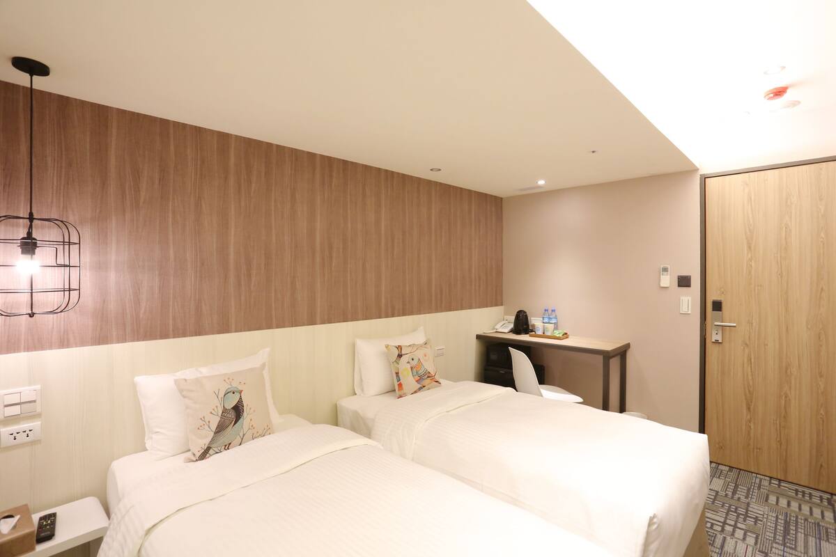 A hotel room with two beds, a desk with a chair, a wooden wall, and a door.