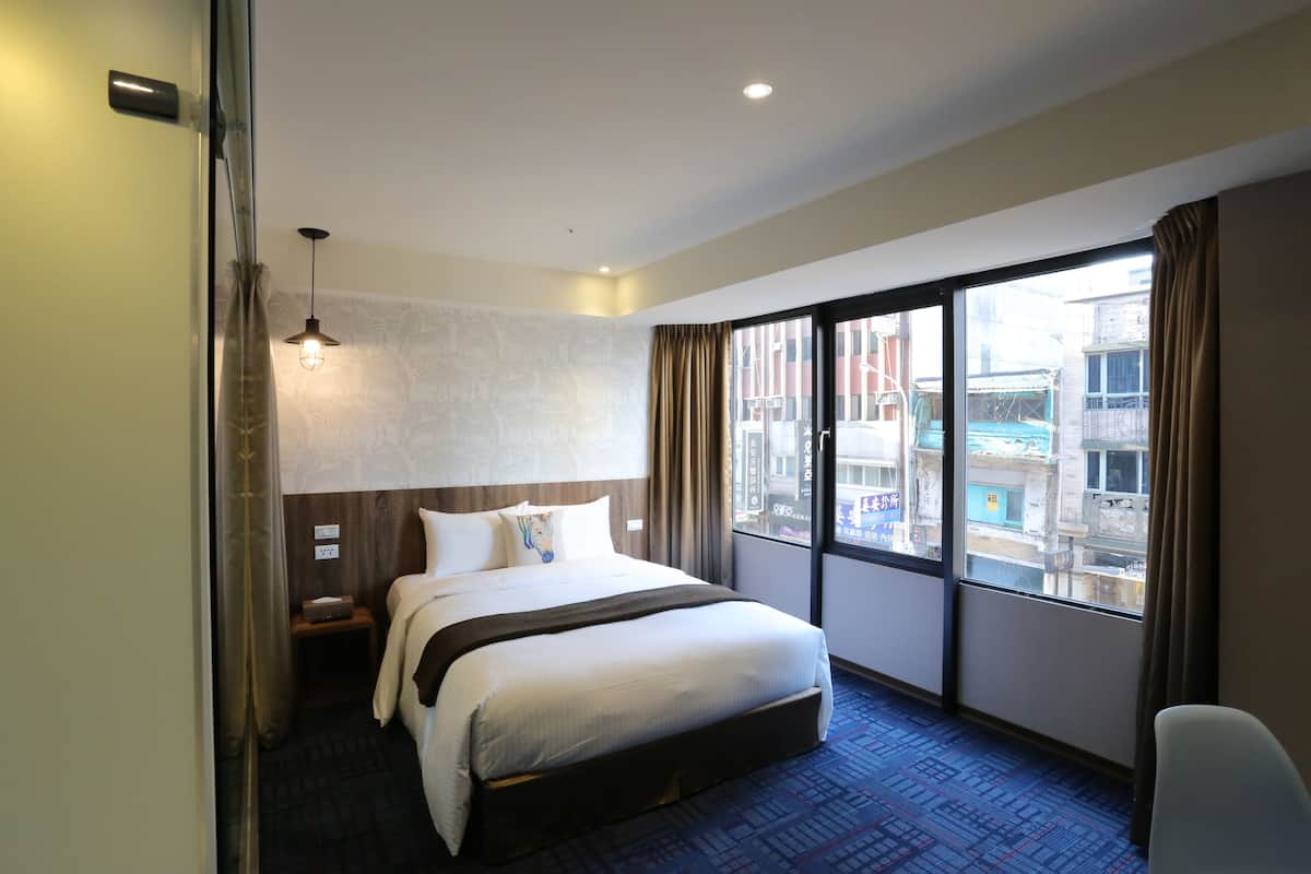 A hotel room with a large bed, a nightstand, a desk, and a window with a city view.