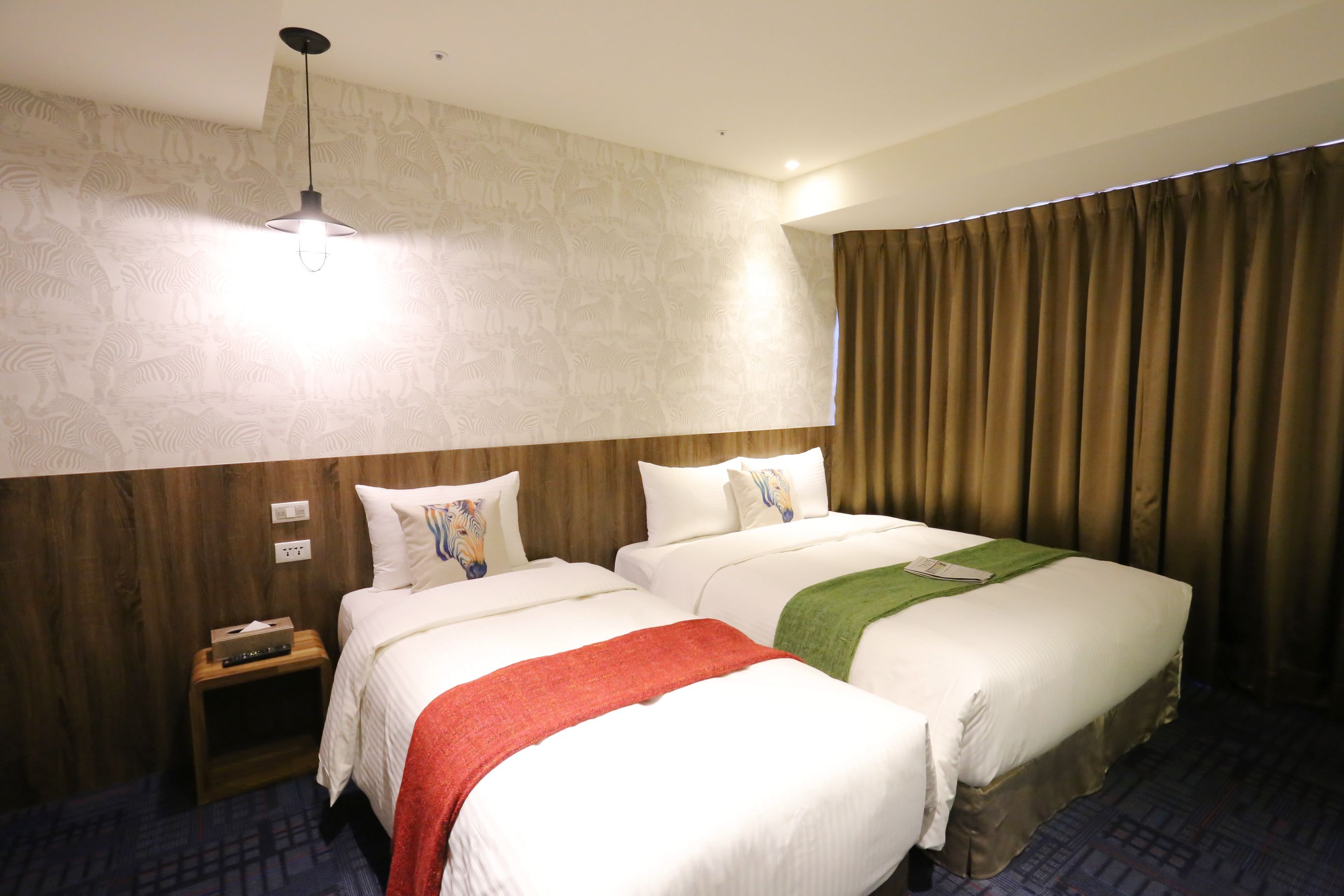 A hotel room with two beds, a nightstand, a lamp, and a curtain.