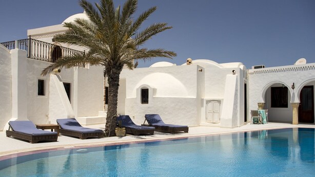 A pool area with lounge chairs and a palm tree, surrounded by white buildings with arches and a clear blue sky. - Maison Leila (Mezraia)
