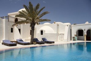 A pool area with lounge chairs and a palm tree, surrounded by white buildings with arches and a clear blue sky. - Maison Leila (Mezraia)