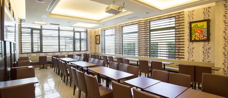 A spacious dining area with multiple tables and chairs, large windows with blinds, and a ceiling projector.