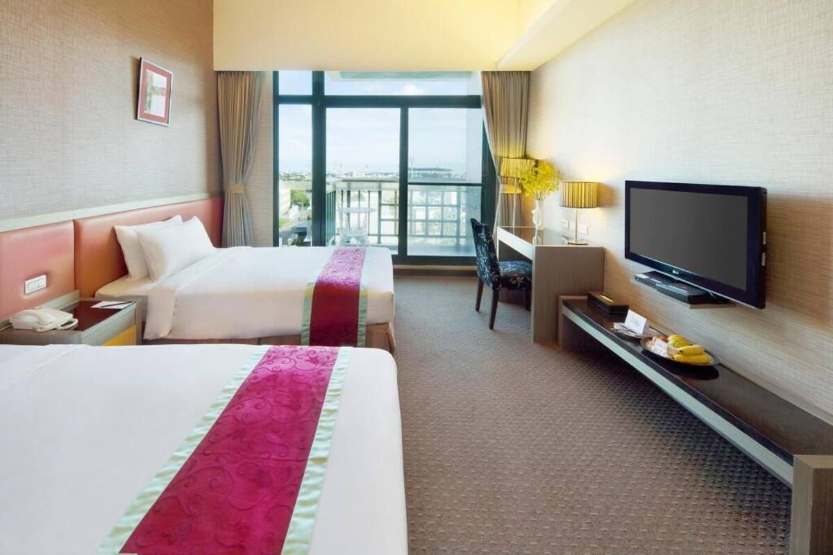 Habitació d'hotel amb dos llits, una televisió, escriptori amb làmpada i vista a la ciutat a través de grans finestres.
