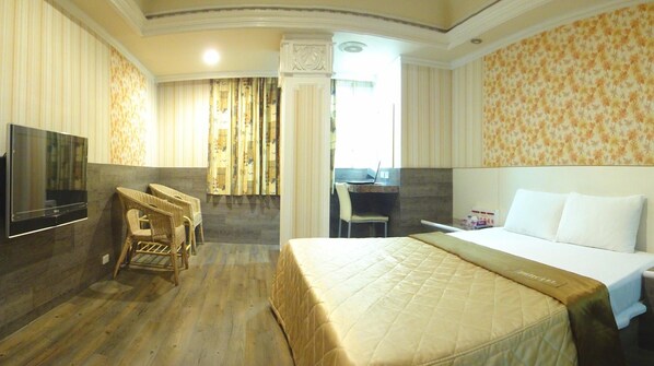 A hotel room with a large bed, a desk with a laptop, a television, and wicker chairs.