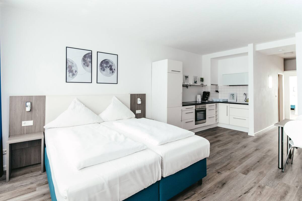 A modern bedroom with a large bed, a small desk, and a kitchenette in the background.
