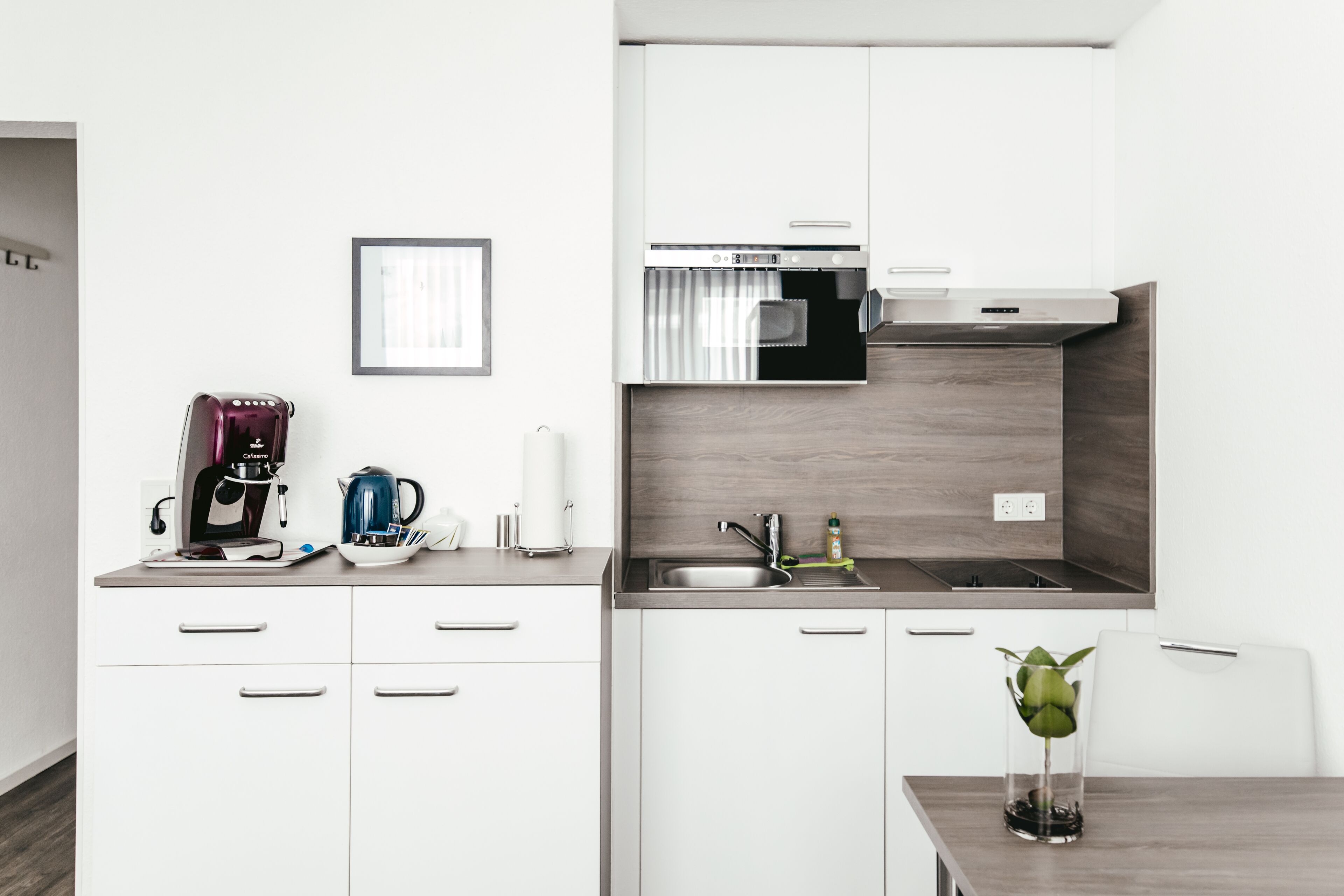 A modern kitchen with white cabinets, a built-in oven, and a sink. There is a coffee maker, a kettle, and a glass vase with a plant on the counter.