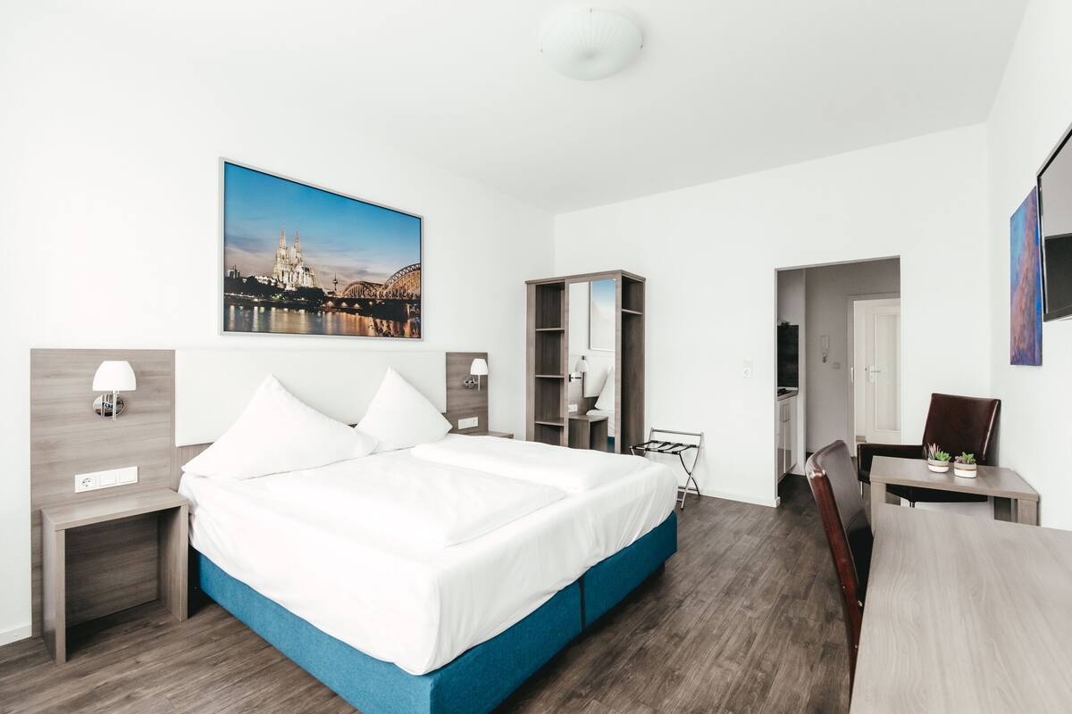 A hotel room with a large bed, a desk, a chair, a wardrobe, and a picture of a cathedral on the wall.