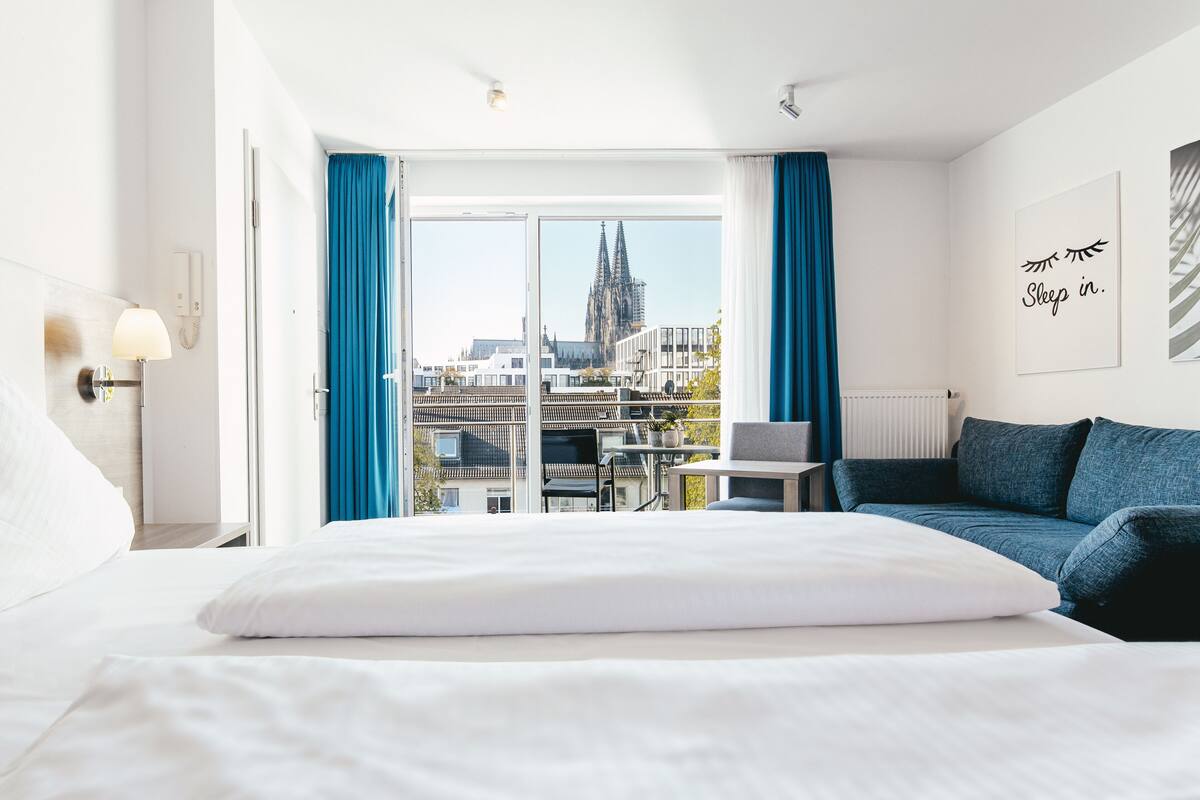A hotel room with a large bed, a desk, a blue sofa, and a view of a cathedral through the window.