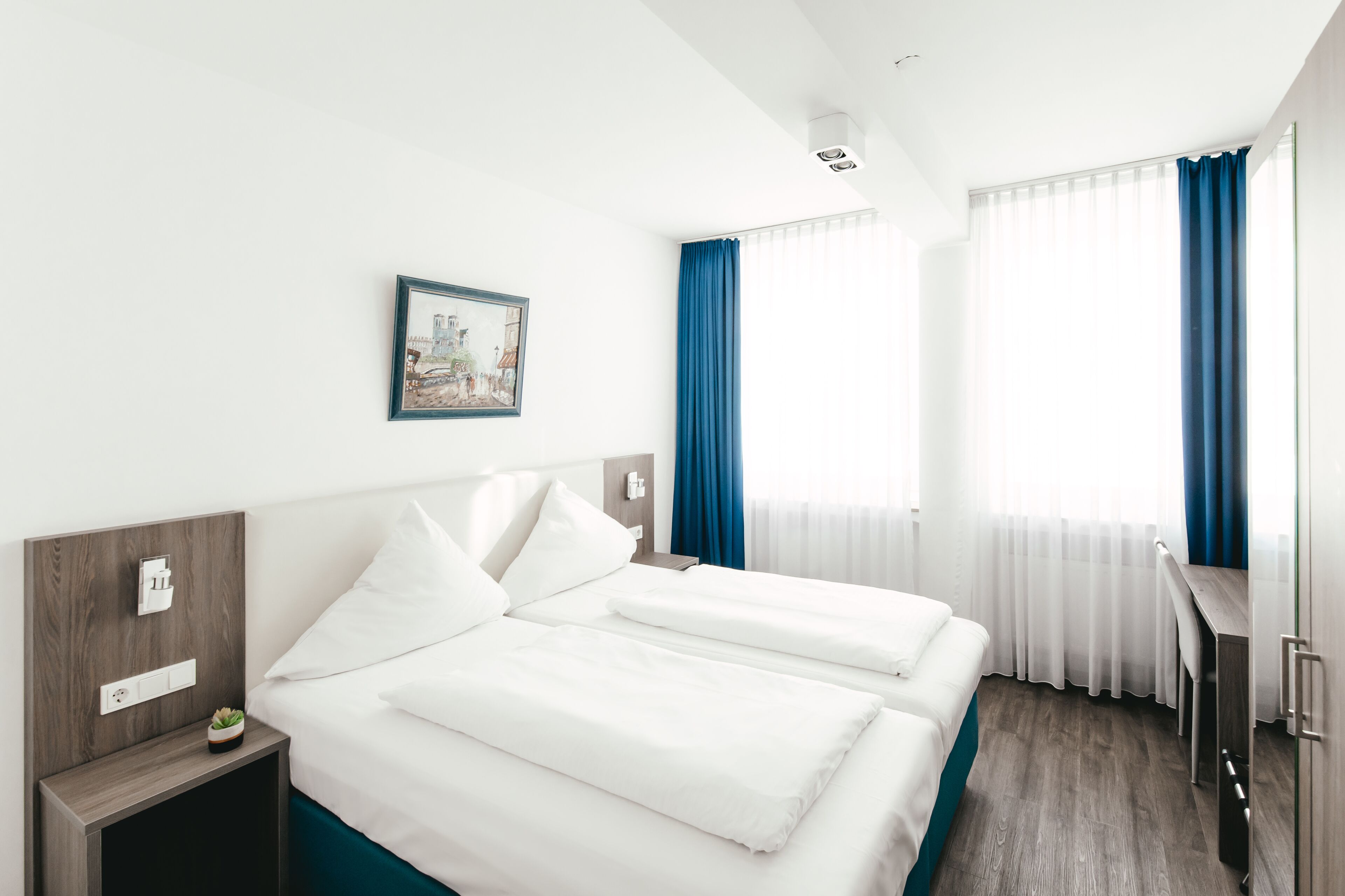 A hotel room with two beds, a desk, and a chair. There is a painting on the wall, a window with curtains, and a small plant on the nightstand.