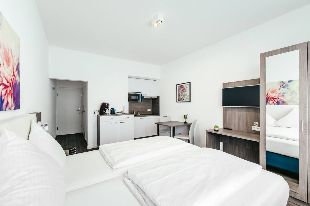 A modern hotel room with a large bed, a desk, a kitchenette, and a TV.