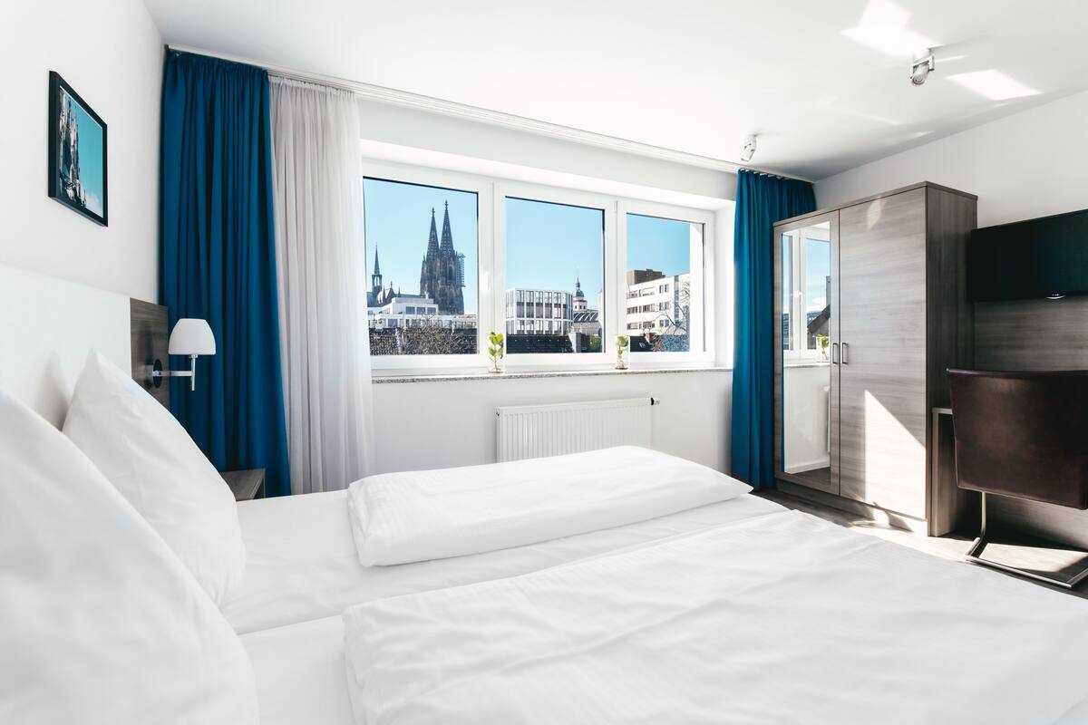 A hotel room with a large bed, a wardrobe, a desk, and a window with a city view.