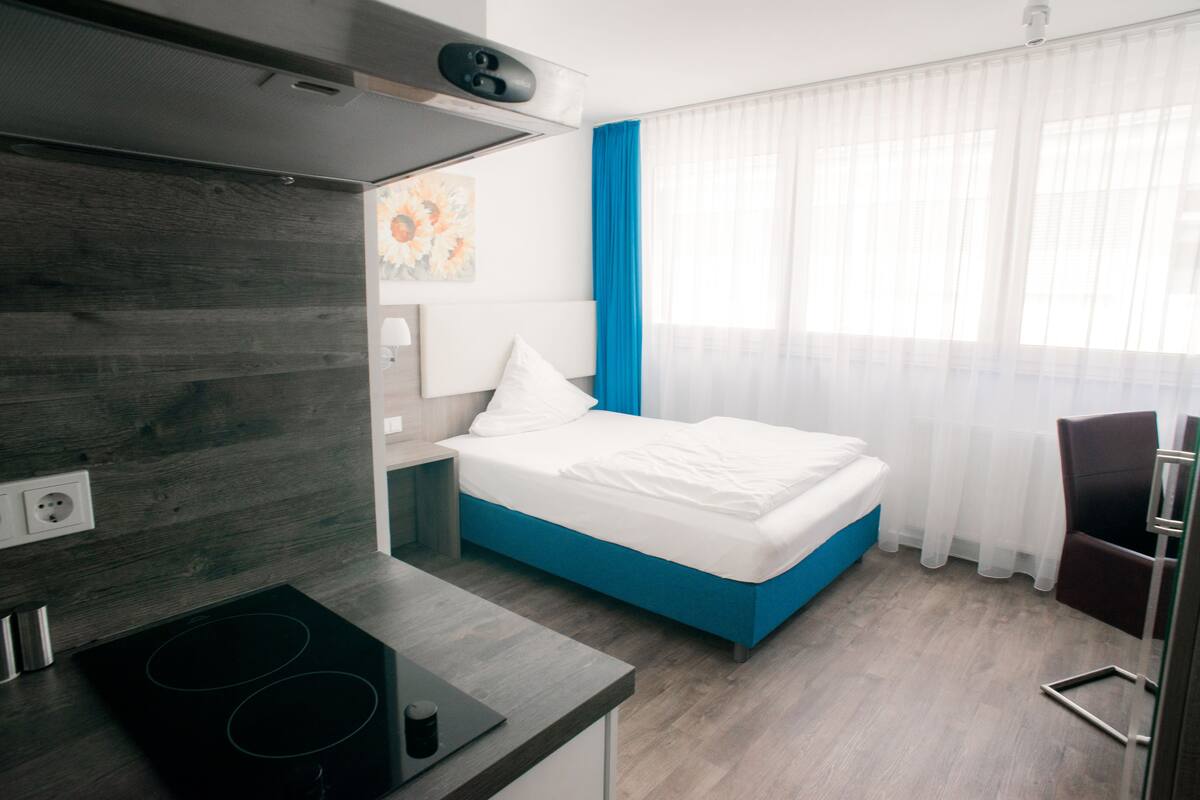 A modern hotel room with a bed, a kitchenette, and a window with sheer curtains.