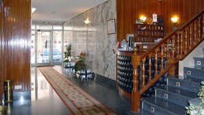 A hotel lobby with a marble wall, a wooden staircase, and a reception desk. - Hotel América (Ferrol)