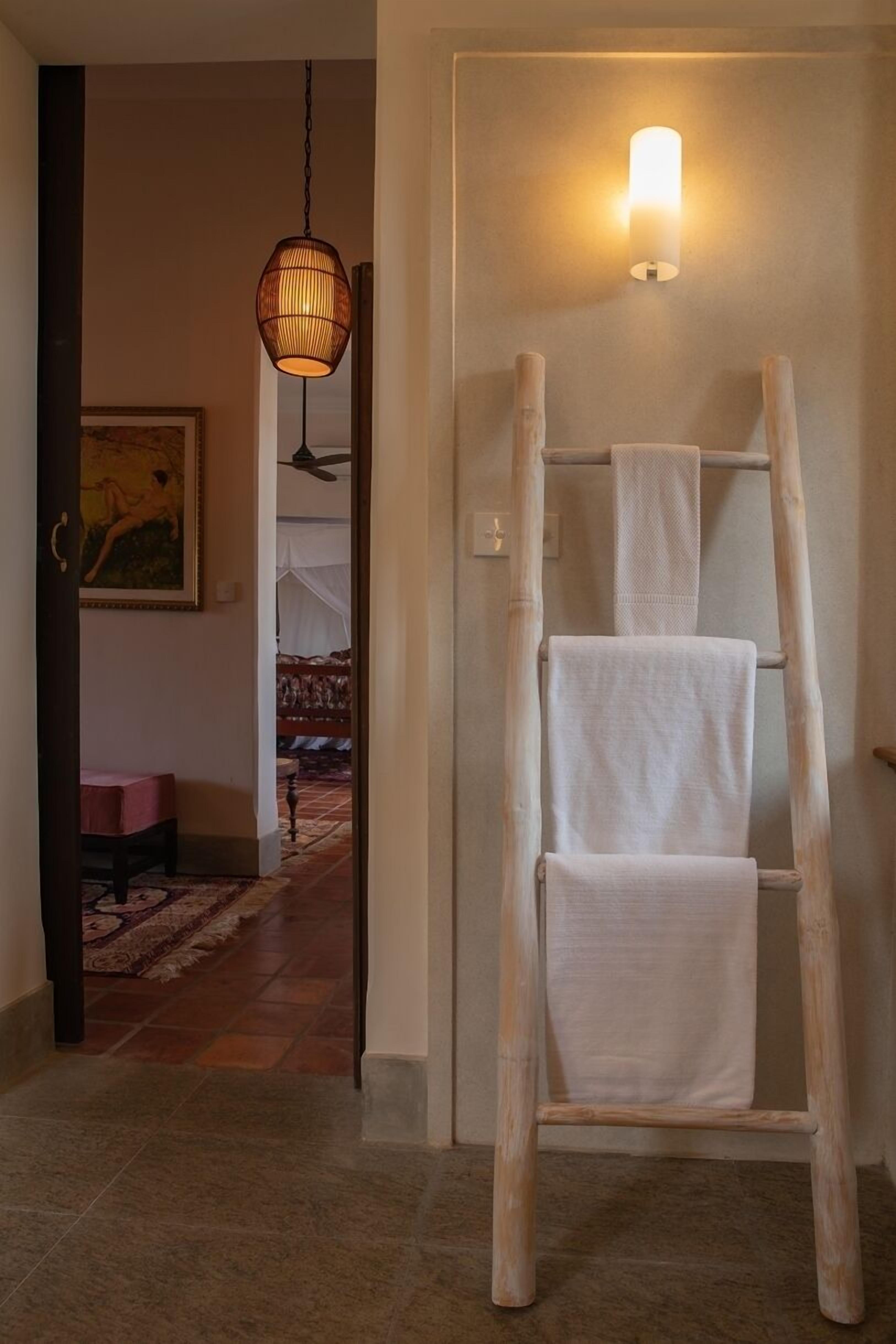 A wooden ladder with towels, a hanging light fixture, and a doorway leading to a room with a bed and a painting.