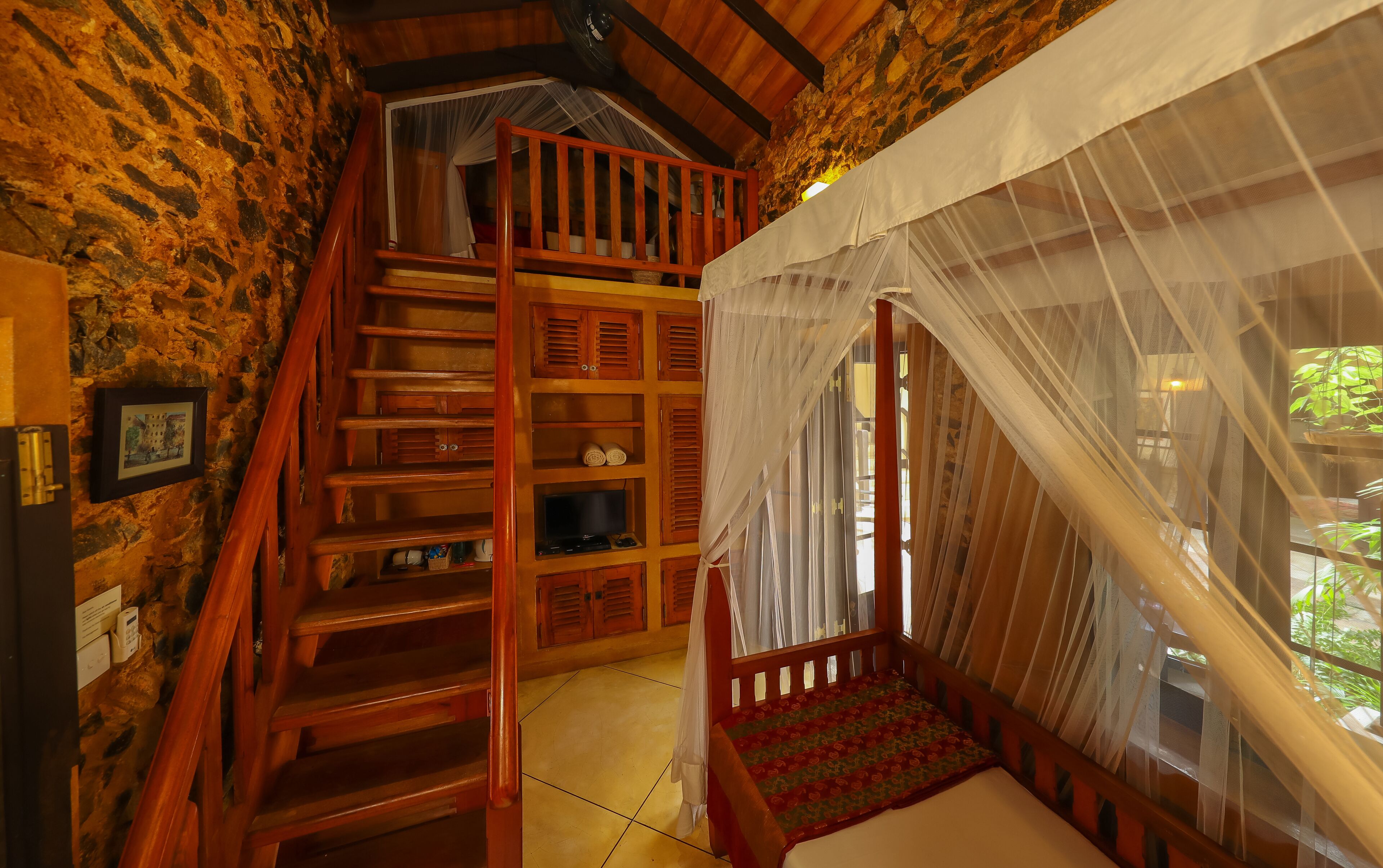 Eine Holztreppe führt zu einem Schlafzimmer mit Himmelbett, Steinwand und Blick auf Grün.