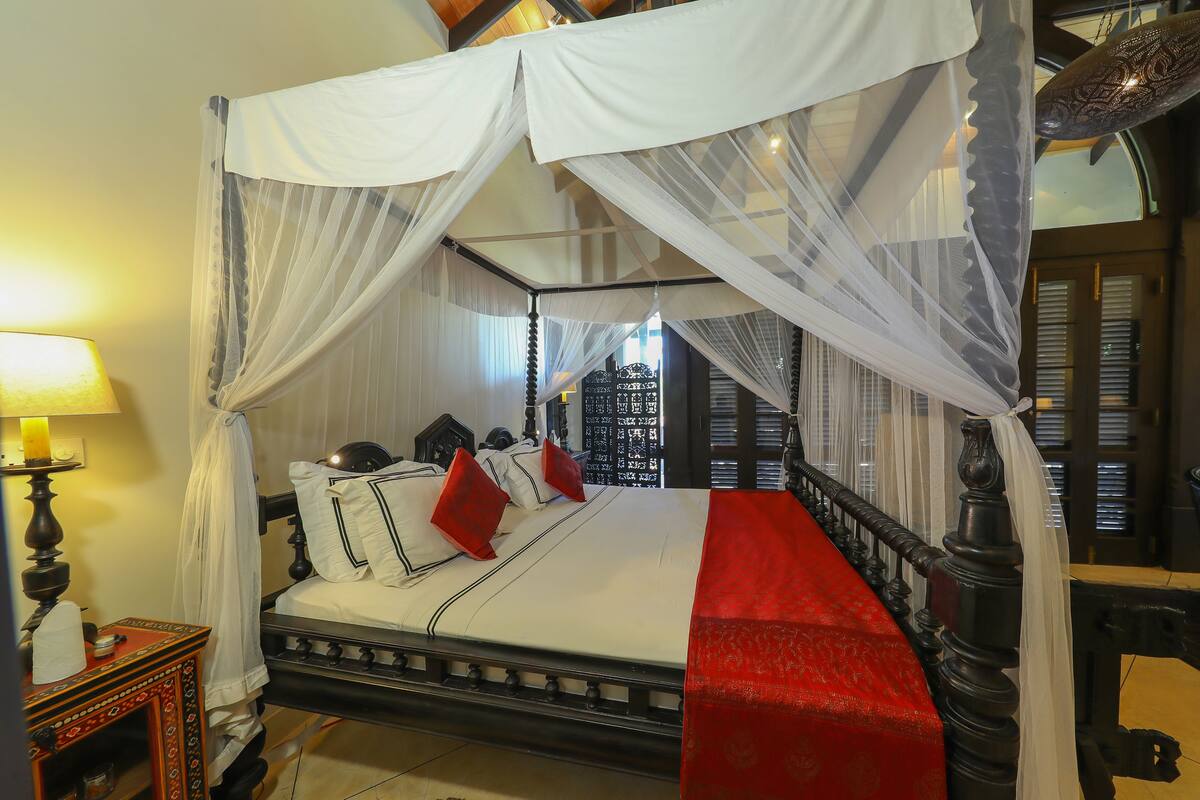 A four-poster canopy bed with white bedding and red pillows, a red runner, and a nightstand with a lamp.