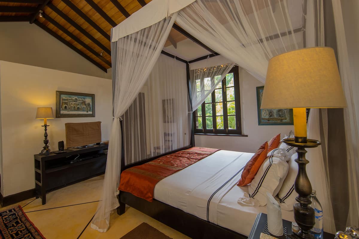 A bedroom with a four-poster bed, a desk with a lamp, and a window with curtains.